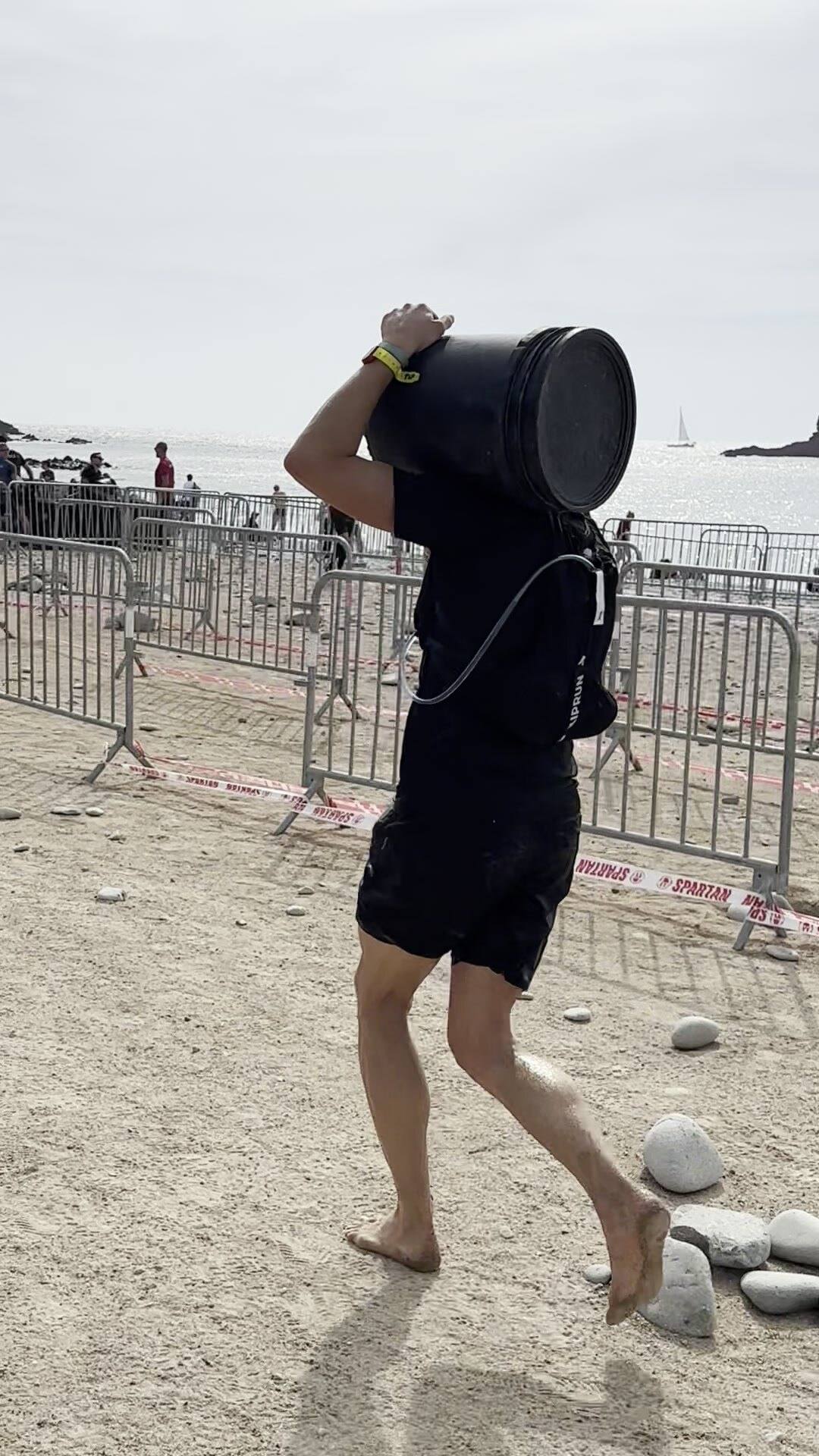 Antoine porte pieds nus un seau noir de gravier sur l’épaule, sur une plage de sable et de galets