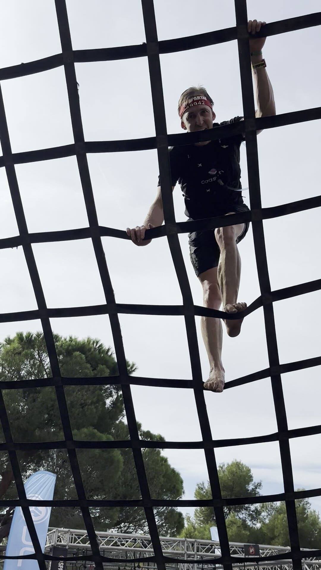 Antoine grimpe pieds nus un filet vertical, orteils enroulés autour d’une corde