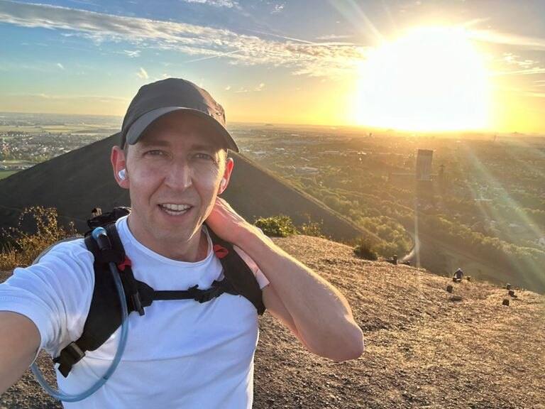 Selfie d’Antoine en casquette et sac d’hydratation au sommet d’un terril, soleil rasant derrière lui